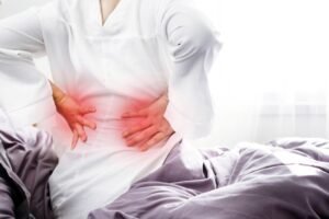 person waking up holding lower back in pain in bedroom morning sunlight